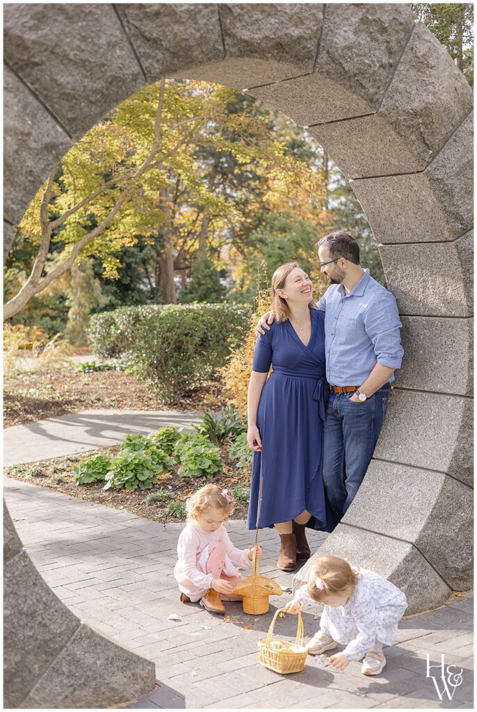 Parents smiling beneath a stone arch while their children play nearby during an outdoor family pictures